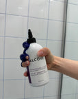 Hand holding a bottle of Alcôve Violet Shampoo in a shower