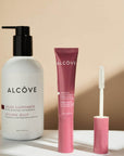 Three Alcôve hair styling products arranged on a white surface against a warm beige background: a large white bottle with a black pump labeled Styling Jelly, a pink squeeze tube labeled Brow/Forehead Brow Product, and a matching pink applicator wand.