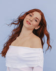 Woman with long red hair wearing a white off-shoulder top against a light blue background