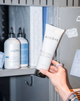 Hand holding a tube of Alcove skincare product in front of a cabinet with more Alcove products.