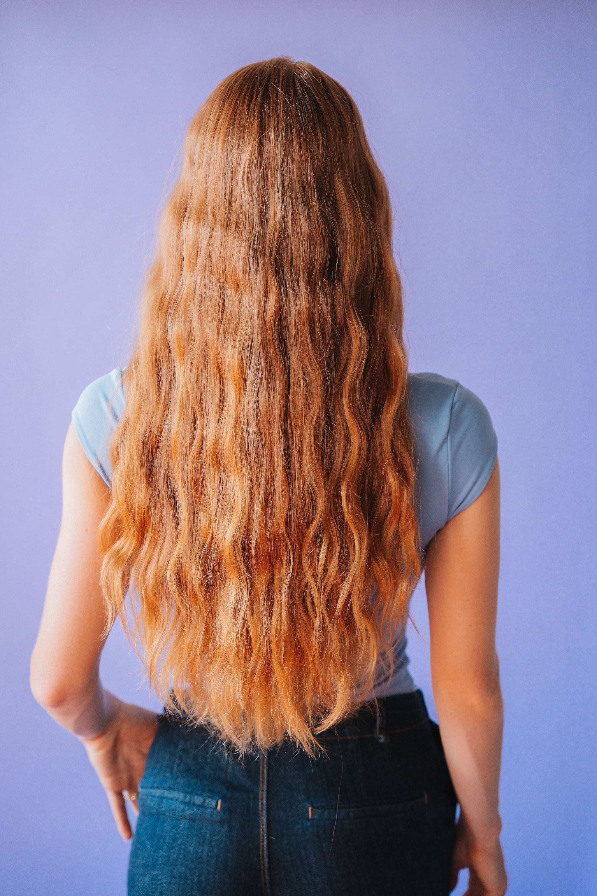 Person with long, wavy blonde hair standing against a purple background