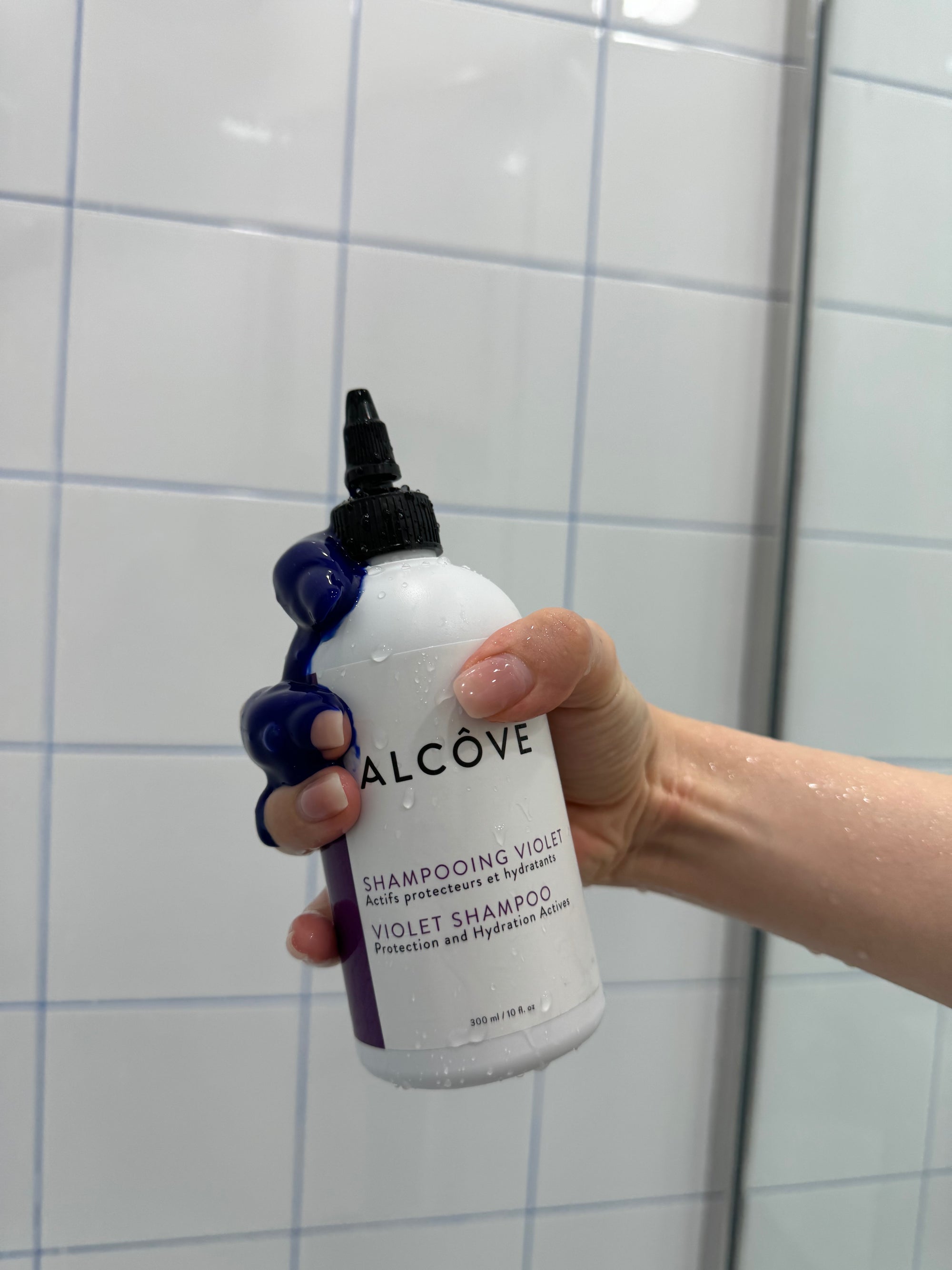 Hand holding a bottle of Alcôve Violet Shampoo in a shower