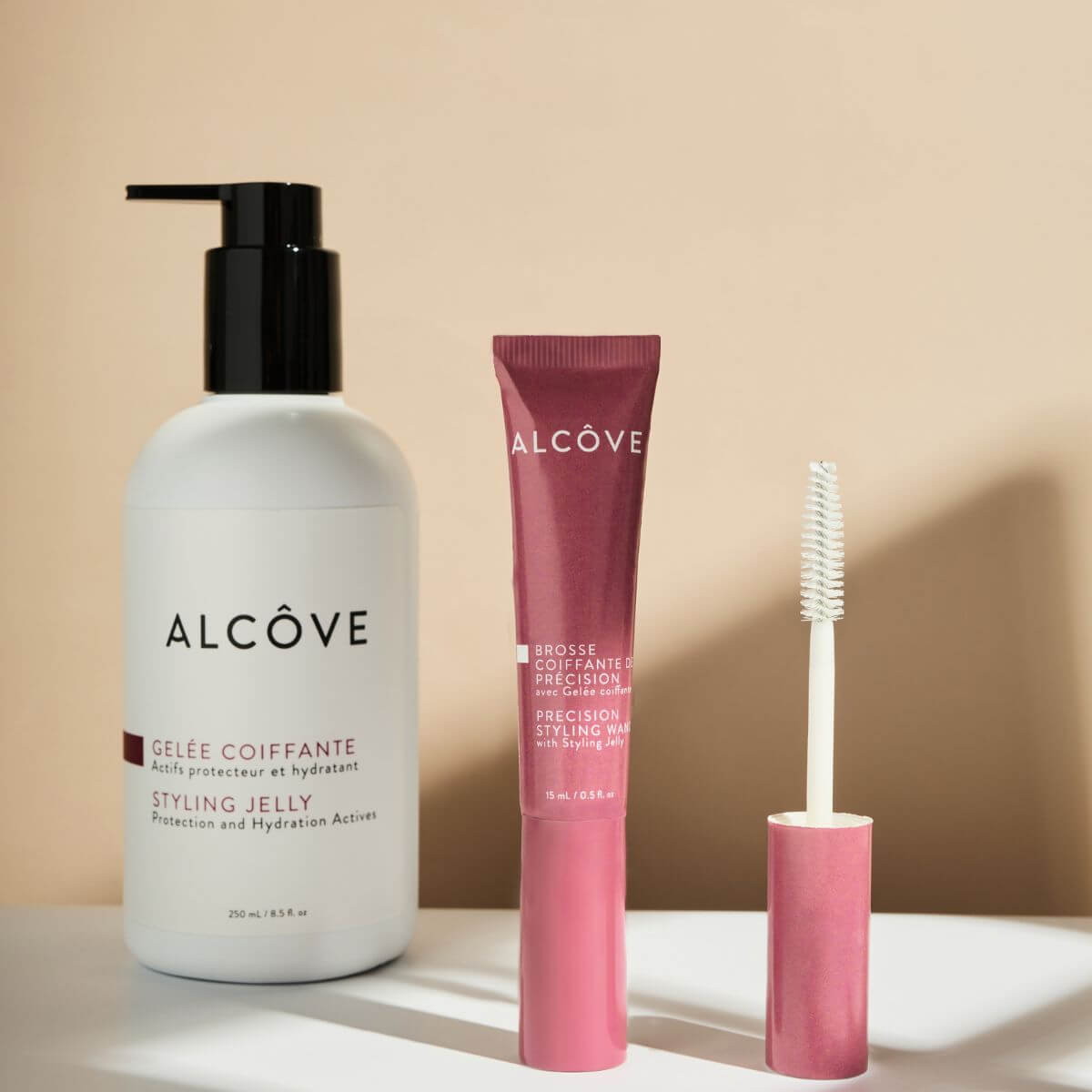 Three Alcôve hair styling products arranged on a white surface against a warm beige background: a large white bottle with a black pump labeled Styling Jelly, a pink squeeze tube labeled Brow/Forehead Brow Product, and a matching pink applicator wand.