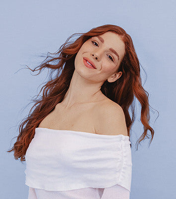 Woman with long red hair wearing a white off-shoulder top against a light blue background
