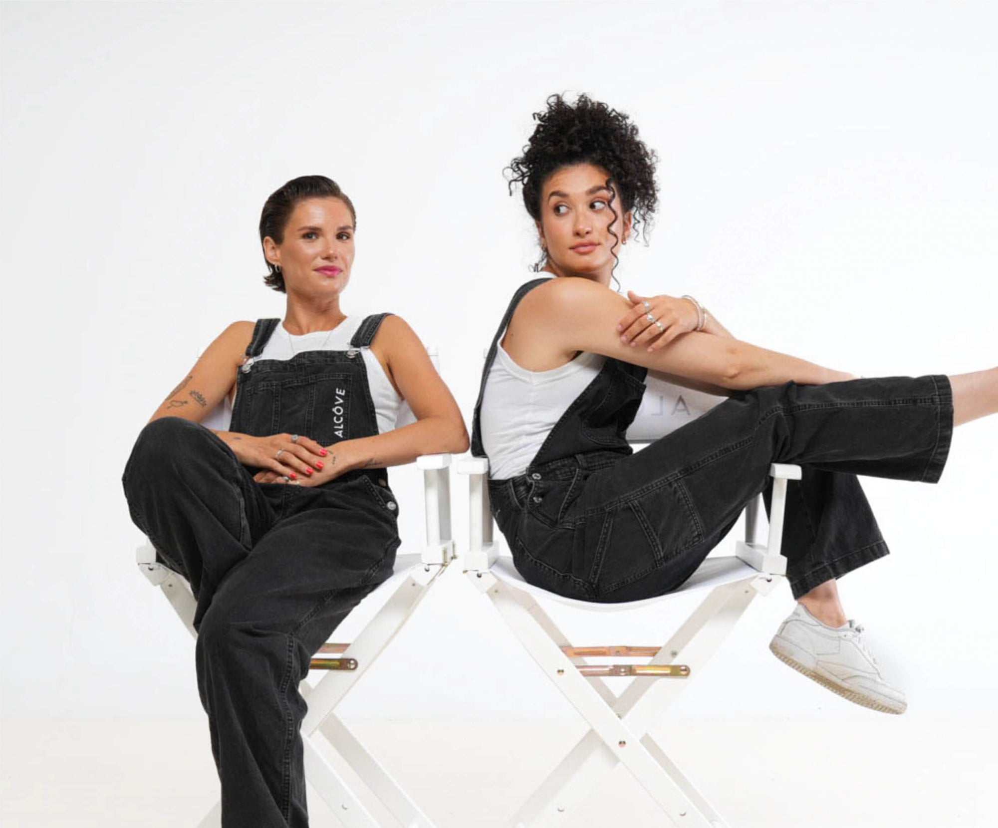 Two women wearing black overalls sitting on white chairs against a white background