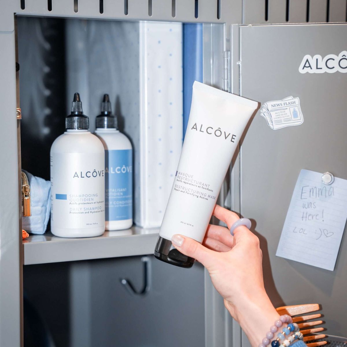Hand holding a tube of Alcove skincare product in front of a cabinet with more Alcove products.