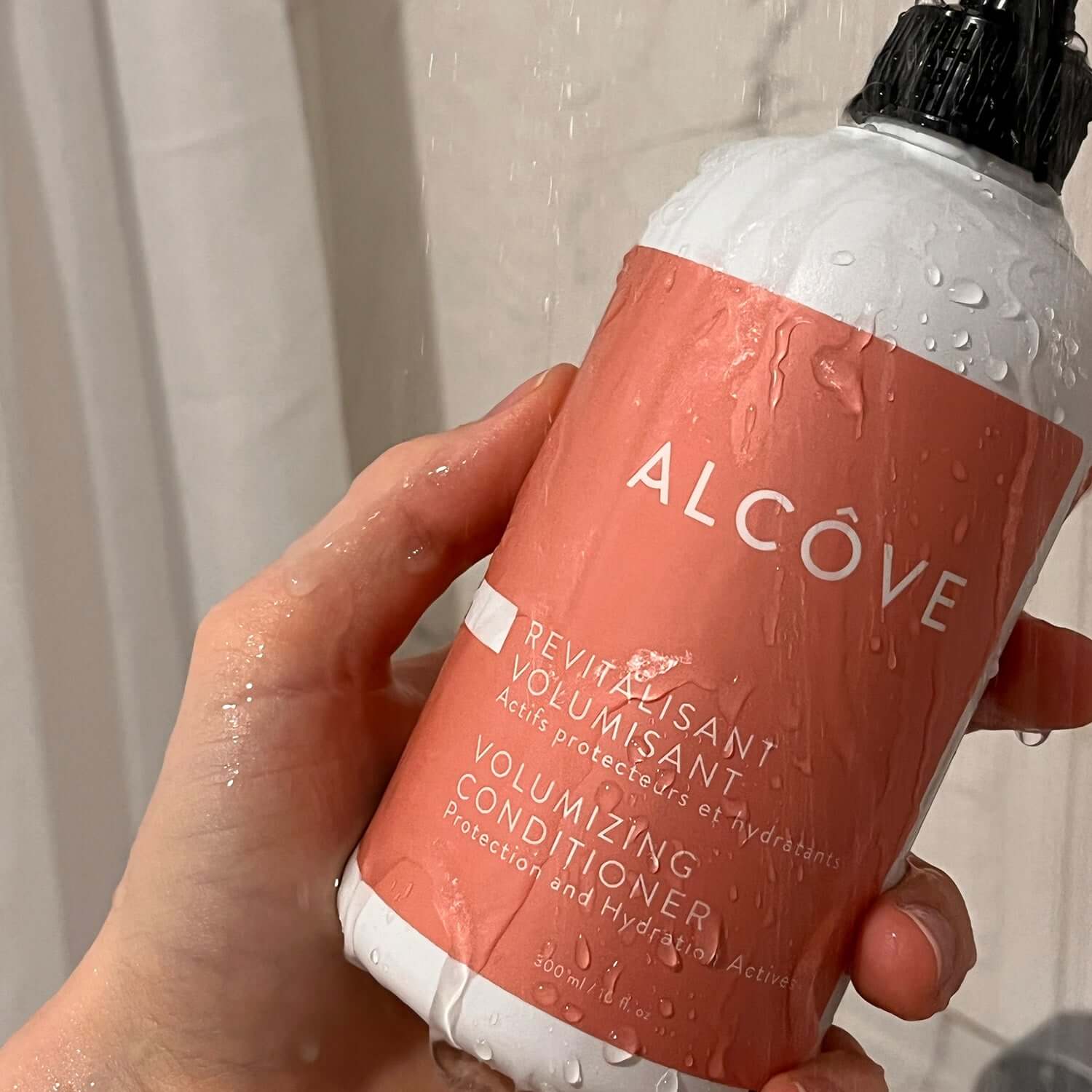Hand holding a bottle of Alcove Volumizing Conditioner with water droplets on the label.